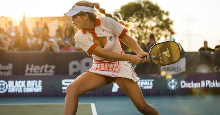 A female athlete aggressively swings a paddle on a pickleball court during sunset, with spectators in the background.